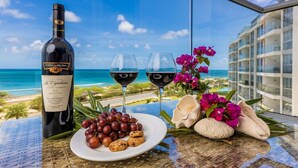 A bottle of La Capitana Merlot, two glasses of red wine, a plate with grapes and cookies, flowers, and a view of the ocean and hotel buildings.