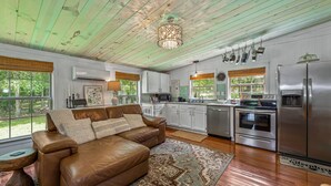 A spacious living room with a brown leather sofa, a patterned rug, and a large window. The kitchen features white cabinets, stainless steel appliances, and a hanging pot rack. - Mountain Meadow (Mentone)