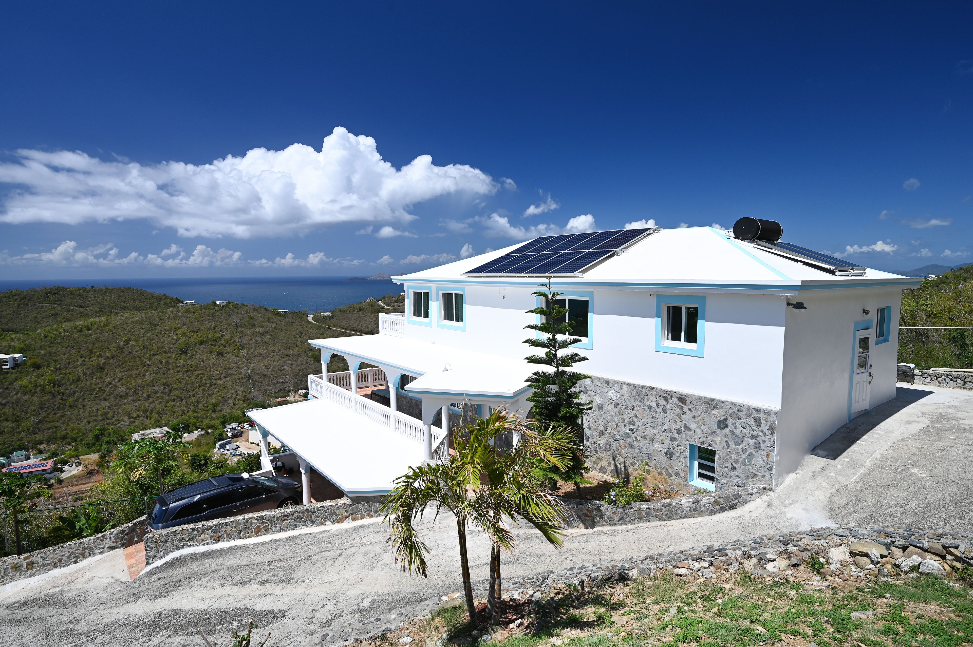 Una casa bianca con tetto blu e pannelli solari, circondata da un muro in pietra e una palma.