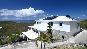Una casa bianca con tetto blu e pannelli solari, circondata da un muro in pietra e una palma.