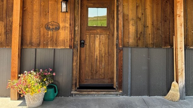 A wooden building with a sign reading "BUNKHOUSE" above a door. There are string lights above the door, and a potted plant with flowers is visible to the left. - Bunkhouse with Horse Accommodations (Big Timber)