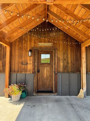 Un bâtiment en bois avec une enseigne indiquant « BUNKHOUSE » au-dessus d’une porte. Des guirlandes lumineuses sont suspendues au-dessus de la porte, et un pot de fleurs est visible sur la gauche.