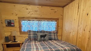 A wooden cabin bedroom with a plaid bedspread, a window with a stained glass design, and a small nightstand with a lamp. - Bunkhouse with Horse Accommodations (Big Timber)