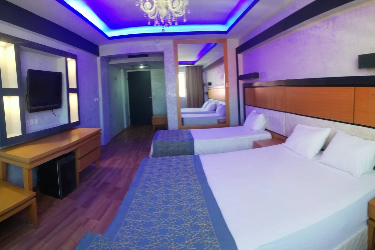 A hotel room with a large bed, a smaller bed, a desk, a TV, and a chandelier.