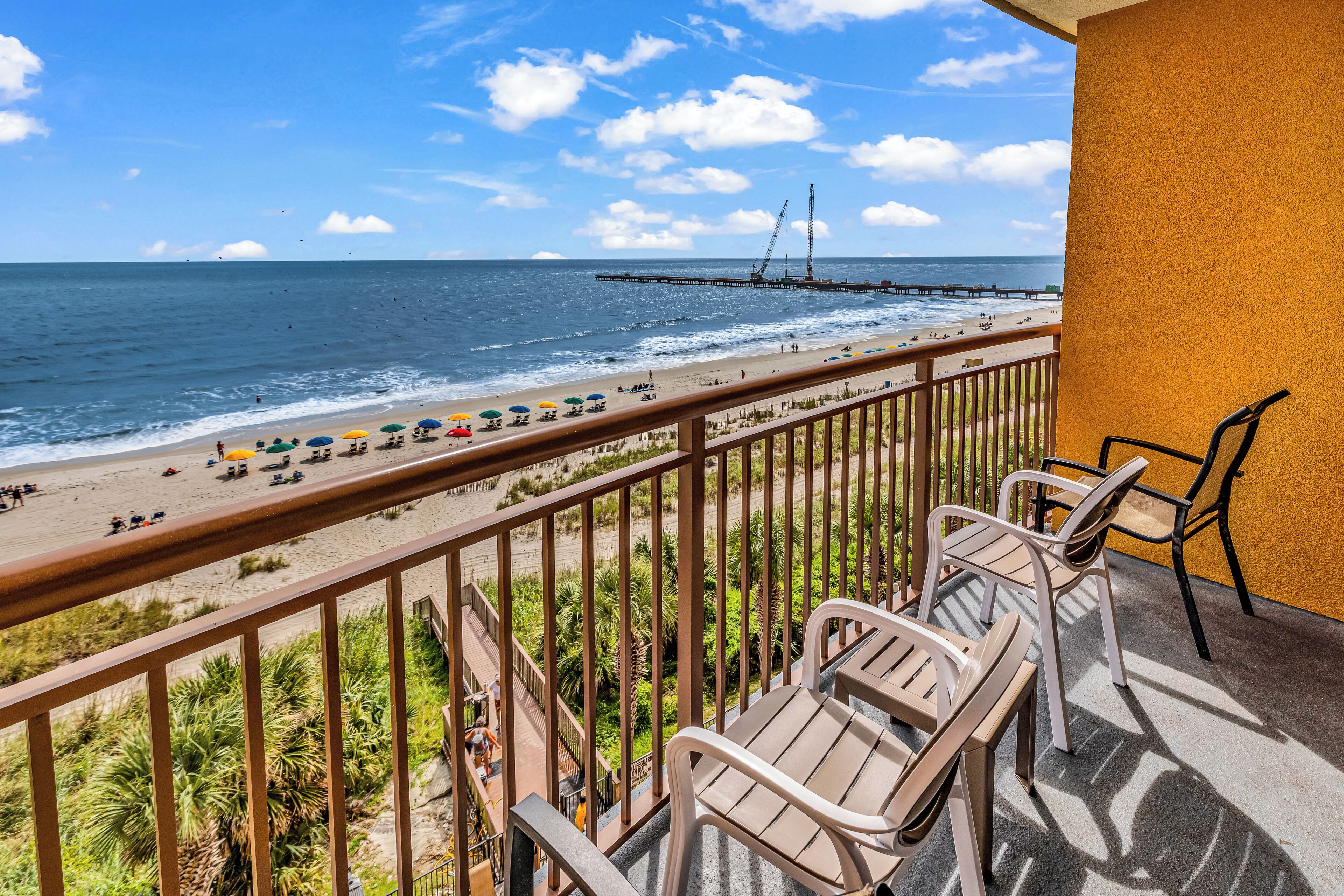 A balcony with a view of the beach and ocean, featuring a railing, chairs, and a yellow wall.