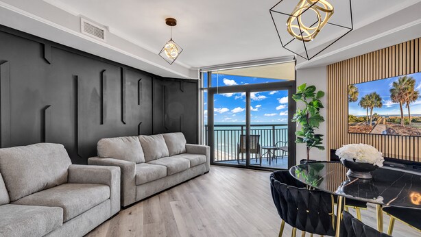 A modern living room with a grey sofa, a glass dining table with black chairs, and a large window with a view of palm trees and a clear sky. - Beachfront Anderson Condos by Hosteeva (Myrtle Beach)