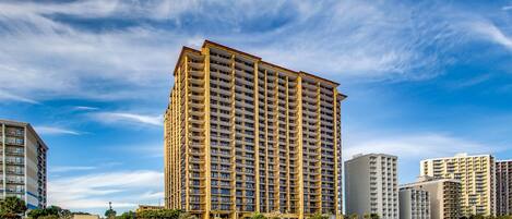 A high-rise hotel with multiple balconies, situated near a beach.