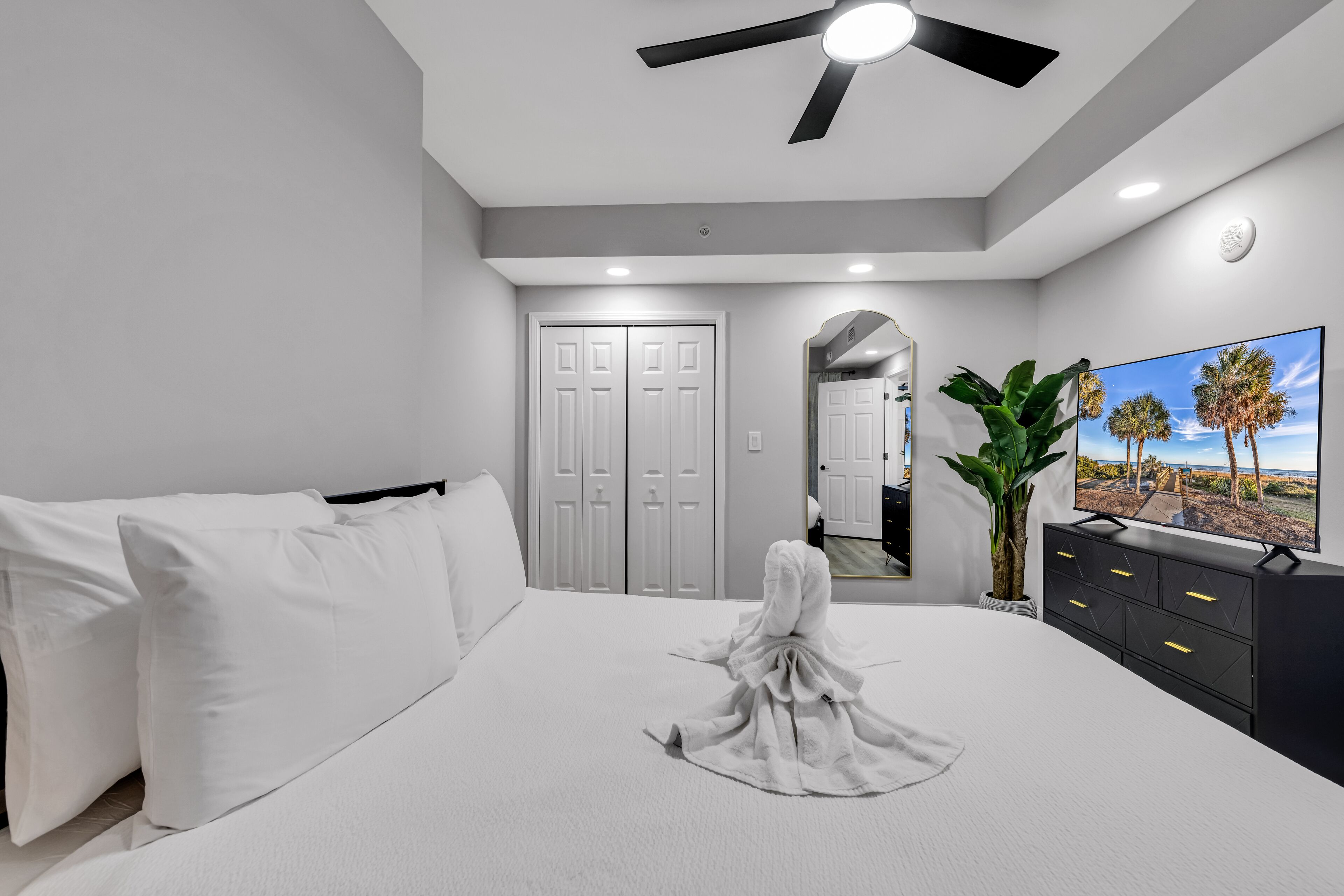 A bedroom with a bed, white pillows, a robe, a ceiling fan, a TV, a plant, and a door.