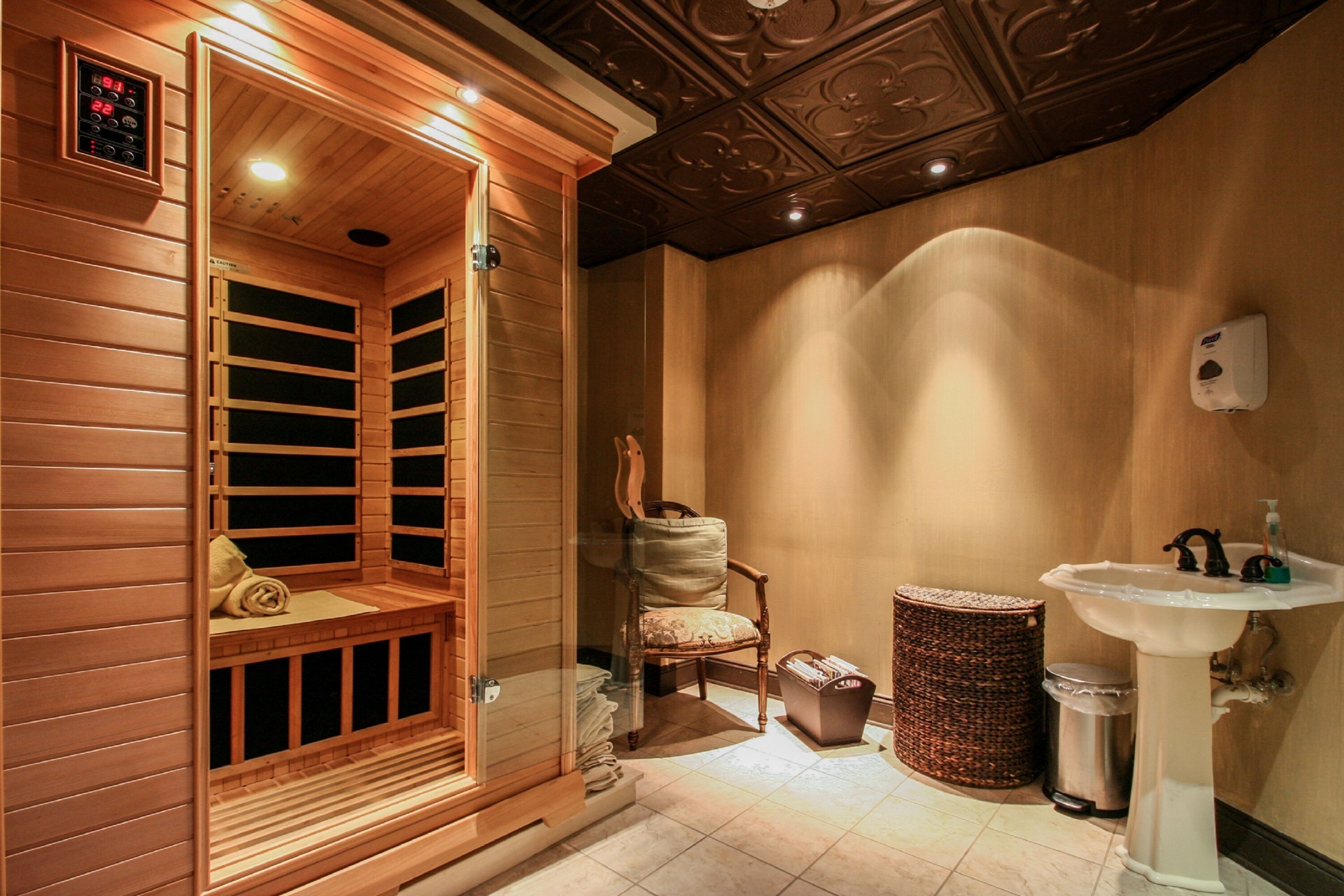 A sauna with wooden walls and a bench, a chair, a wicker basket, a trash bin, and a sink with a mirror.