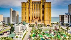 A large yellow hotel with multiple floors, a parking lot, and a pool area with palm trees. - Beachfront Anderson Condos by Hosteeva (Myrtle Beach)