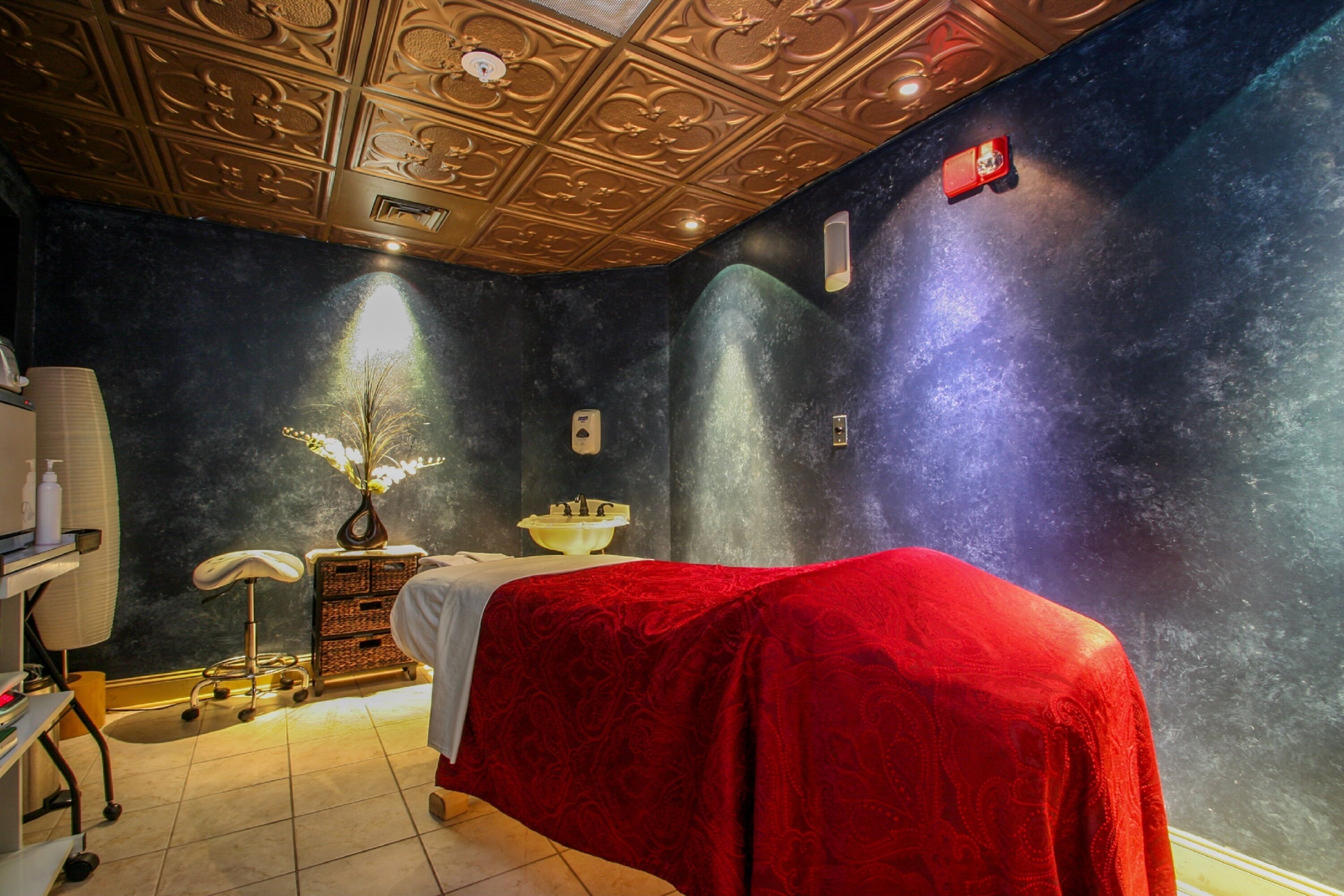 A spa room with a massage table covered in red, a small stool, a white basin, and a decorative plant.