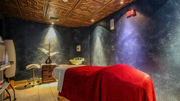 A spa room with a massage table covered in red, a small stool, a white basin, and a decorative plant.