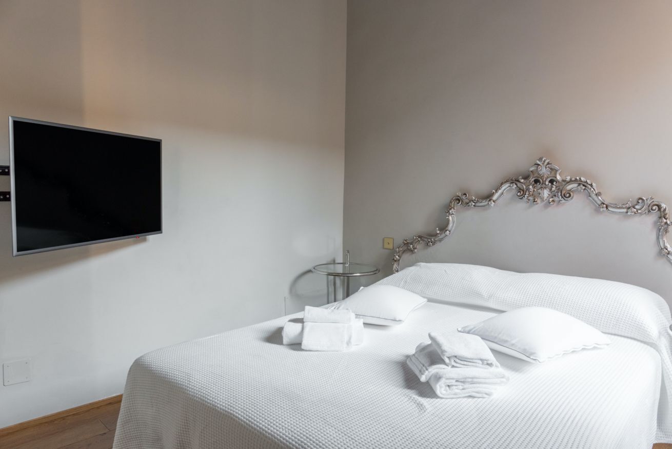Un lit bien fait, avec du linge de lit et des serviettes blanches, un téléviseur à écran plat fixé au mur et une tête de lit décorative.