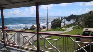 Une vue sur la côte, depuis un espace abrité doté d’une balustrade, donnant sur l’océan et une route menant à une plage bordée de rochers et de palmiers.