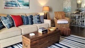 A living room with a sofa, coffee table, and a wicker chair. There is a lamp, a painting, and a dining area in the background. - Island House D : Stylish Updated Townhome - Steps to the Beach w/ Pool! (Surfside Beach)