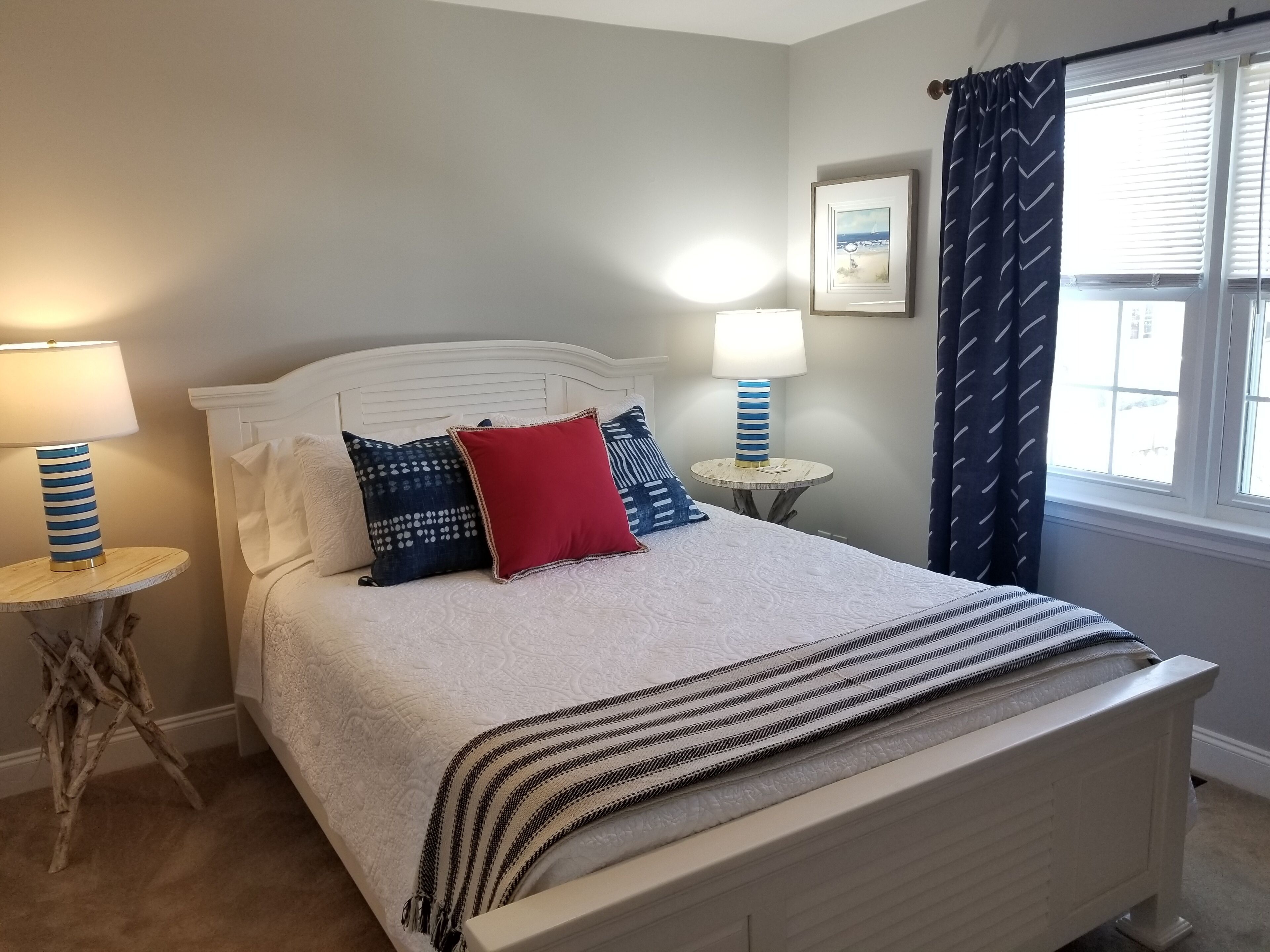 A neatly made bed with a white headboard, two bedside tables with lamps, a window with curtains, and a framed picture on the wall.