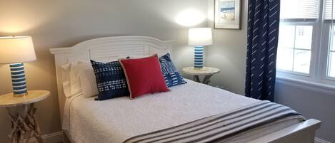 A neatly made bed with a white headboard, two bedside tables with lamps, a window with curtains, and a framed picture on the wall.