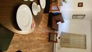 Comedor con mesa de madera preparada para cuatro, con platos, tazas y cubiertos. Hay un sofá marrón y una pequeña mesa auxiliar de madera con un jarrón de flores.