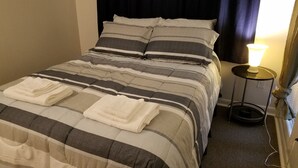 Una cama bien hecha con ropa de cama a rayas y toallas dobladas, una lámpara de noche y una mesita con una lámpara.