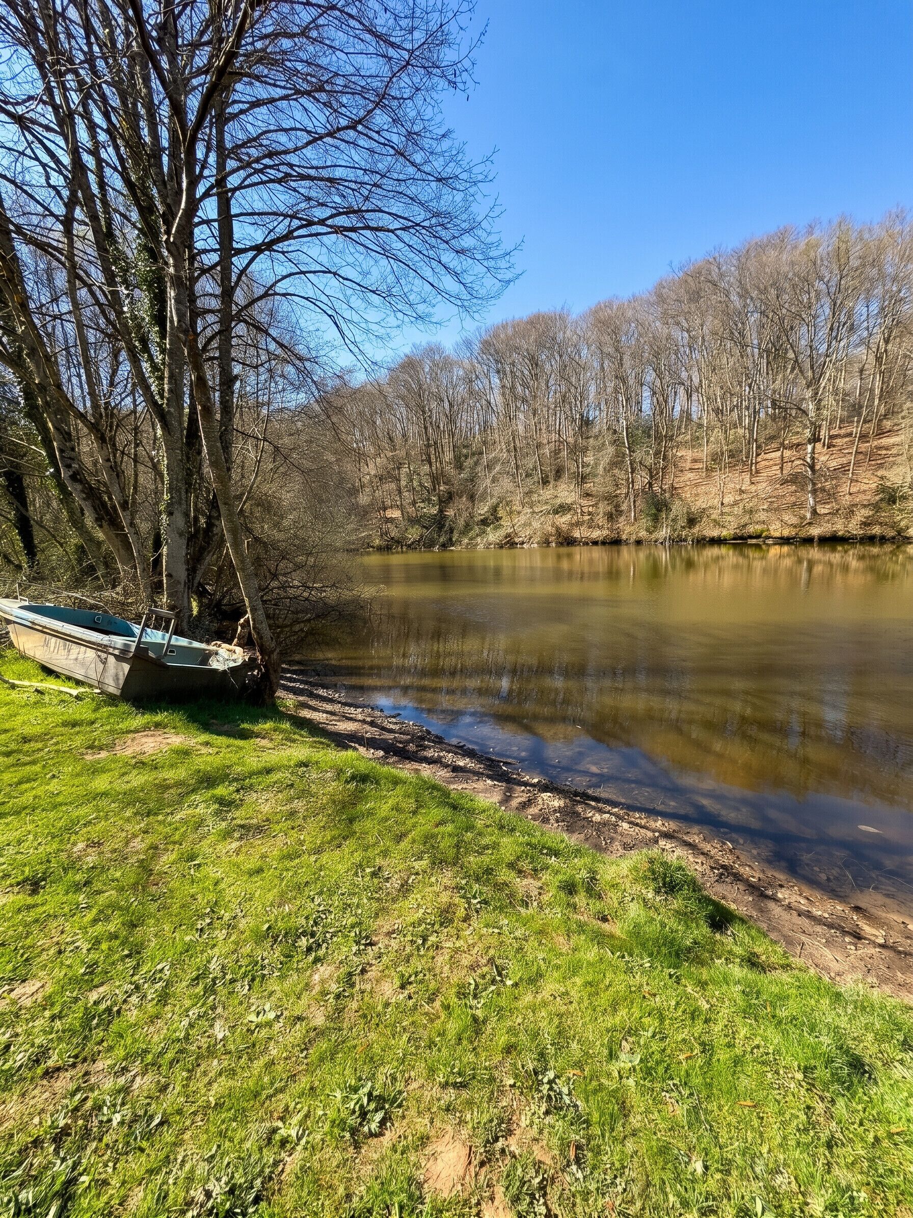 Une rivière paisible, avec un bateau amarré sur la rive gauche, entourée de berges verdoyantes et d’arbres sans feuilles.