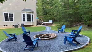 Un jardin avec un foyer entouré de chaises Adirondack bleues, une terrasse en bois et une maison en arrière-plan.