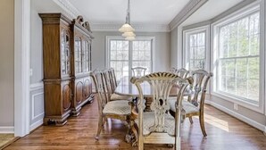 Une salle à manger avec une table en bois et des chaises décorées, de grandes fenêtres et un lustre classique.