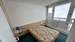 Un dormitorio con cama, mesita de noche, lámpara, ventana y vista a un paisaje nevado, además de un radiador.