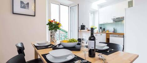 A modern dining area with a wooden table set for two, a vase of flowers, and a bottle of wine.