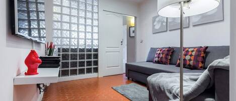 A modern living room with a grey sofa, colorful cushions, a red sculpture, a white shelf, and a large window with a grid pattern.