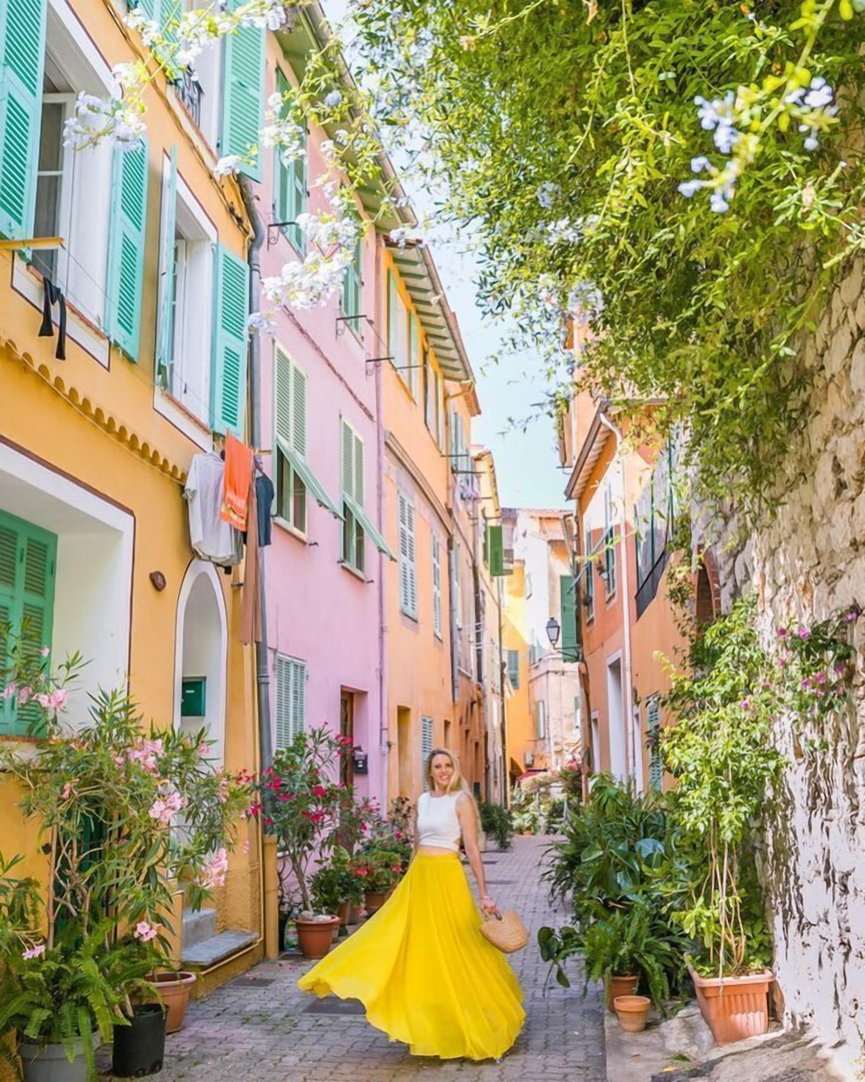 A narrow street with colorful buildings, a woman in a yellow dress, and potted plants.