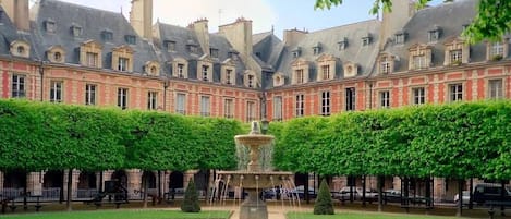 A courtyard with a fountain, surrounded by neatly trimmed hedges and a large building with multiple windows and a red facade.