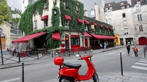 Una scooter roja estacionada en una acera frente a un edificio con toldos rojos y fachada cubierta de hiedra verde.