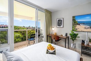Ein Hotelzimmer mit einem Bett, einem Schreibtisch, einem Stuhl, einem Esstisch, einer Topfpflanze und einem großen Fenster mit Blick auf den Strand.