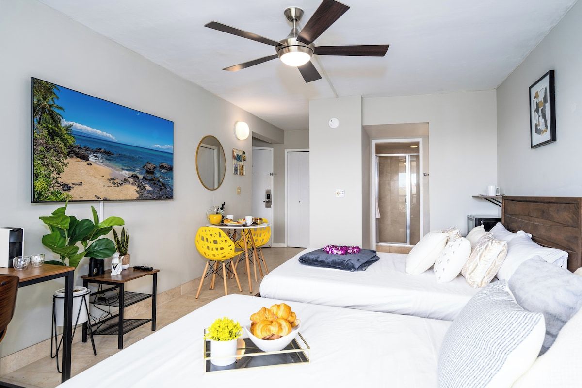 Une chambre d’hôtel comprenant un lit, un bureau, une chaise, une table à manger, un ventilateur de plafond et un tableau représentant une vue sur la plage.