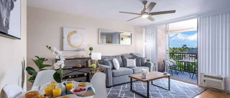 A modern living room with a grey sofa, a dining table with breakfast, a ceiling fan, and a balcony with outdoor furniture.