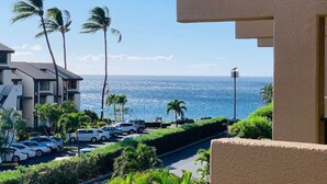 A view of a coastal road with parked cars, palm trees, and a building with a balcony overlooking the ocean. - Luxury Maui Condo | Ocean Views + Resort Perks (Wailea-Makena)