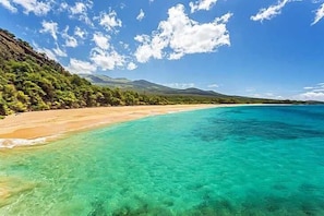 Ein Strand mit klarem türkisblauem Wasser, sandigem Ufer und grünen Hügeln im Hintergrund.