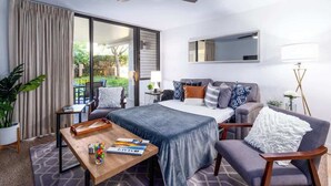 Habitación de hotel con una cama, un escritorio con una bandeja de aperitivos, una silla, un sofá, un ventilador de techo, un ventanal con cortinas y vistas a la vegetación.