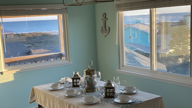 Ein Esstisch gedeckt mit weißem Geschirr und Gläsern, ein Laternenlicht und ein dekoratives Anker an einer taubenfarbenen Wand mit Blick auf das Meer und ein Gebäude.