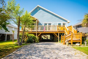A two-story house with a covered porch, a gravel driveway, and a flag. - Pelican Park: Dog-friendly Home Overlooking Canal with Backyard Fireplace, Just (Oak Island)