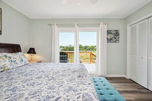 A bedroom with a large bed, a nightstand, a lamp, a view of a deck, and a closet. - Pelican Park: Dog-friendly Home Overlooking Canal with Backyard Fireplace, Just (Oak Island)