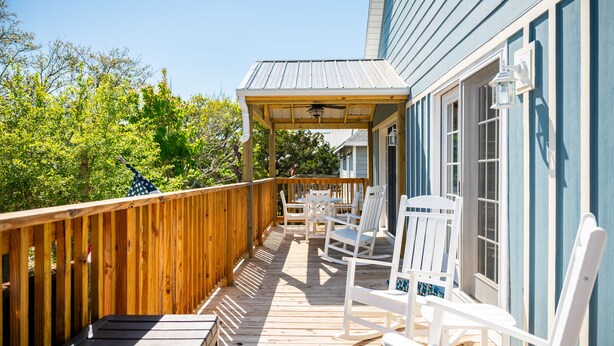 A wooden deck with white rocking chairs and a covered area, surrounded by trees and a house with blue siding. - Pelican Park: Dog-friendly Home Overlooking Canal with Backyard Fireplace, Just (Oak Island)
