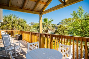 A wooden deck with white rocking chairs and a table, surrounded by palm trees and a clear blue sky. - Pelican Park: Dog-friendly Home Overlooking Canal with Backyard Fireplace, Just (Oak Island)