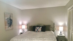 Um quarto com uma cama grande, duas mesas de cabeceira com abajures, um banquinho ao pé da cama e uma obra de arte na parede retratando uma tartaruga.