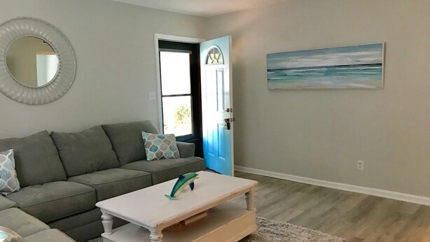 Uma sala de estar com um sofá cinzento, uma mesa de centro branca e um quadro representando uma cena de praia.