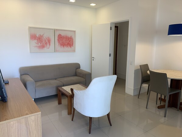 Una sala moderna con un sofá gris, un sillón blanco, una mesa de centro de madera y un comedor con una mesa redonda y sillas.
