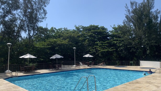 Ein rechteckiger Pool mit Fliesen umgeben, umgeben von Bäumen und beschattet durch Sonnenschirme.
