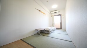 Une pièce avec un sol recouvert de tatamis, un futon servant de lit, une petite table, un climatiseur fixé au mur et une porte coulissante donnant sur un balcon.