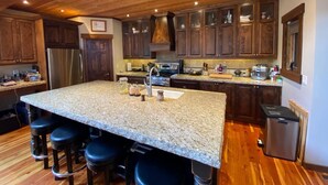 Una cocina con una isla grande, taburetes de bar, armarios de madera, electrodomésticos de acero inoxidable y techo de madera.