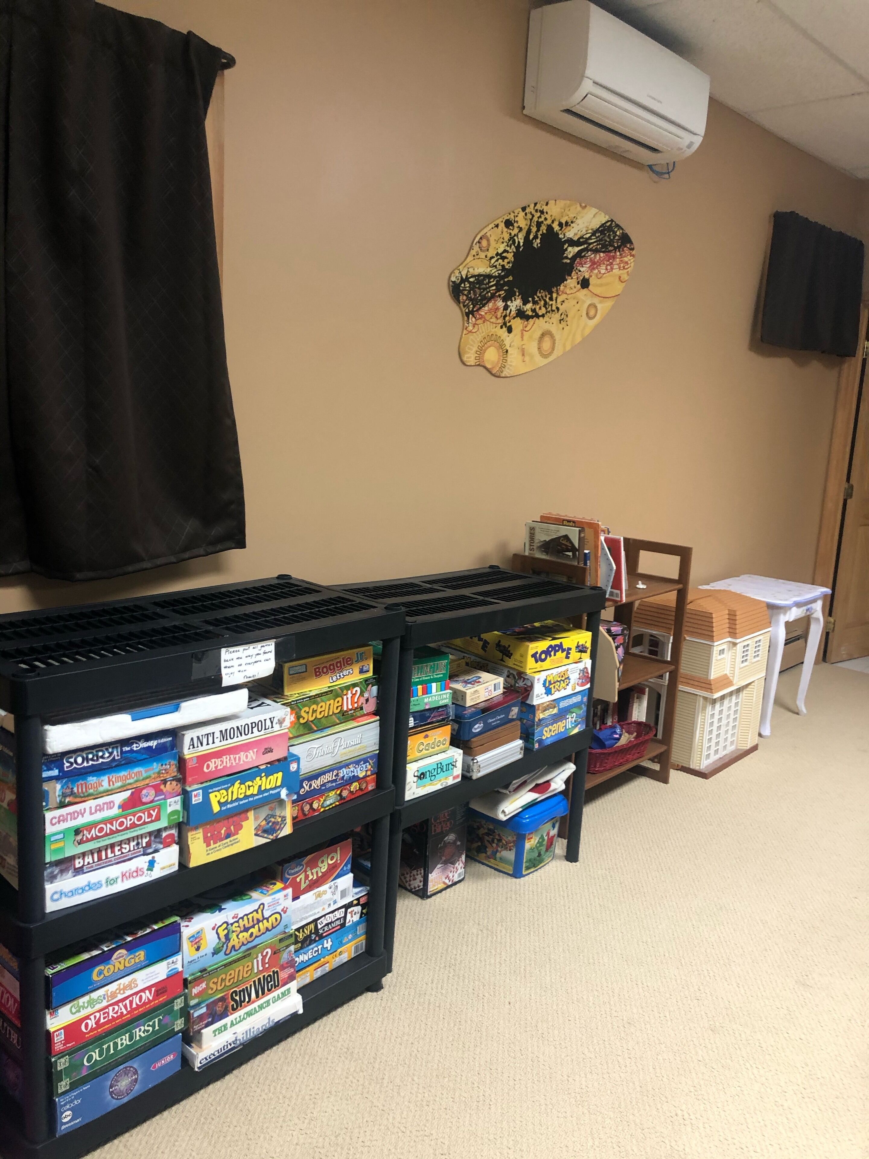 A room with a black shelf filled with board games, a black curtain, a wall-mounted air conditioner, and a small table with a basket.